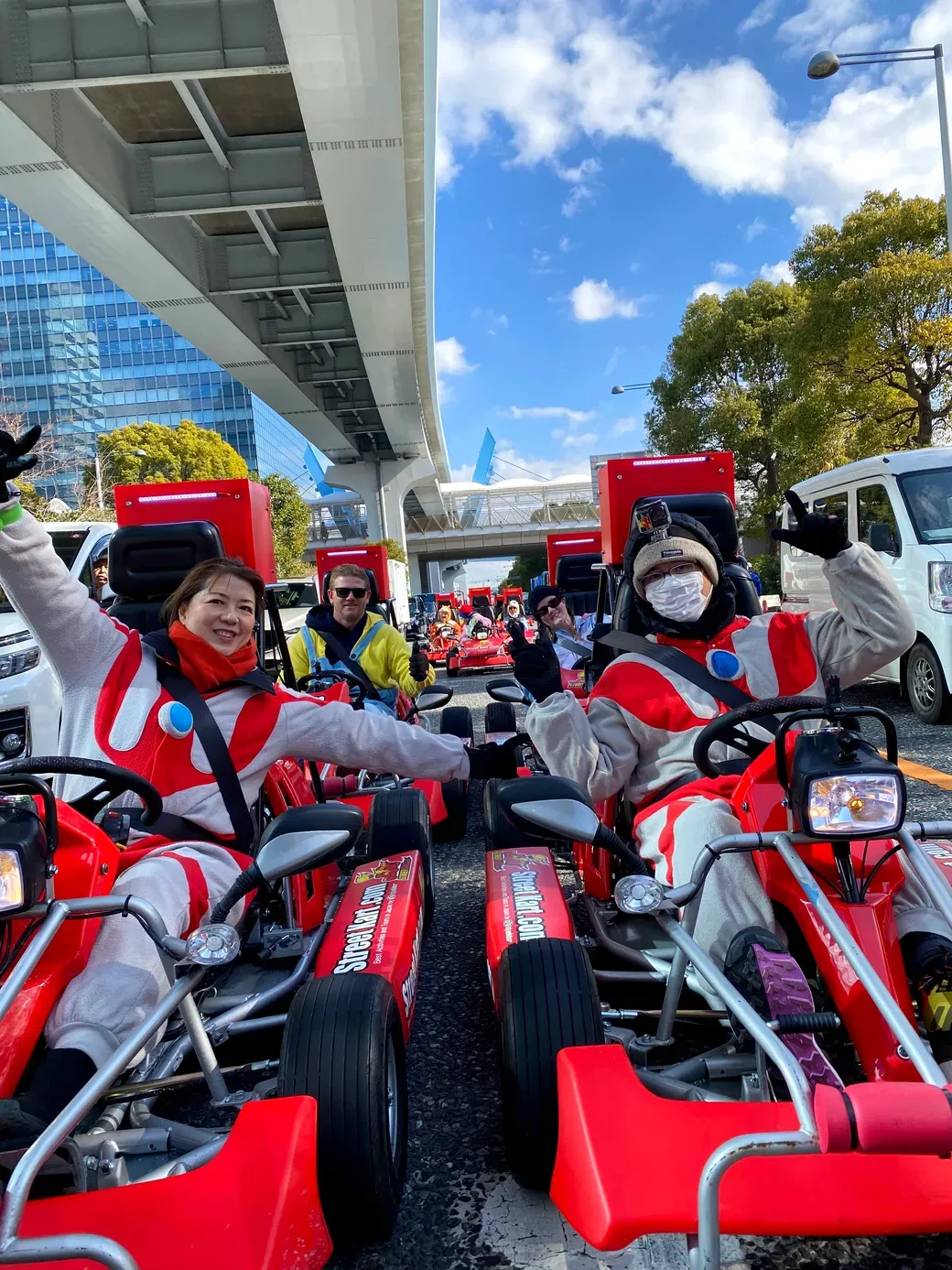 Group of people in red-and-white go-karts posing and smiling under an elevated roadway in a city setting.