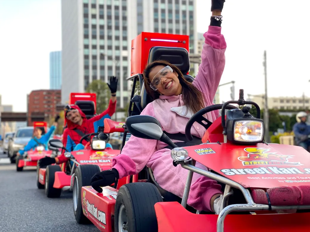 A group of people riding small red go-karts down a city street, smiling and waving with colorful outfits and safety gear.