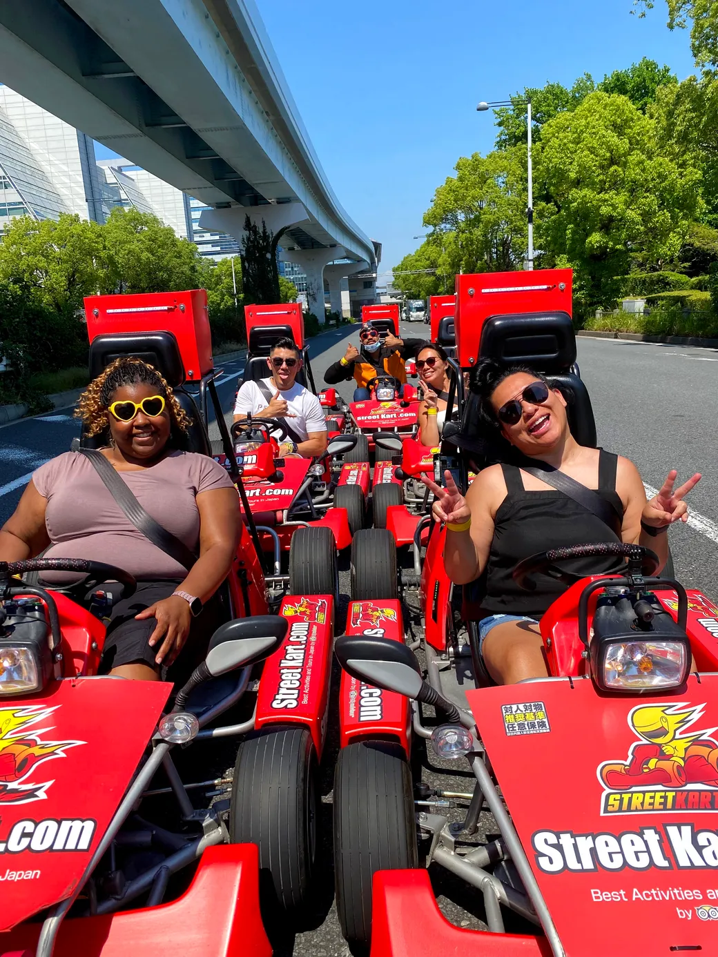 Group of friends riding red go-karts on a sunny street, giving thumbs up and posing for the camera outside urban scenery under a highway overpass.